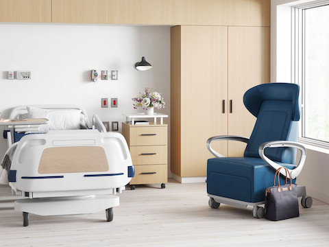 A patient room with the Mirage Overbed Table placed by a hospital bed. A Nemschoff Bedside Cabinet and a Mora wardrobe both in an ash finish next to it, and a Nemschoff Ava Recliner in a blue upholstery nearby.