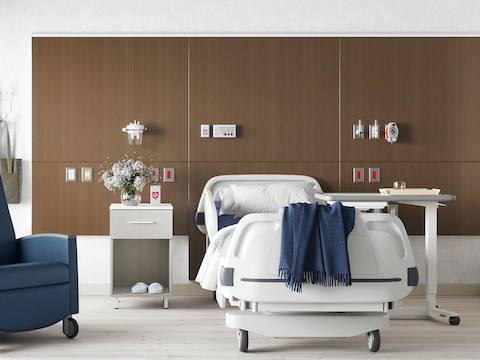 A patient room with a hospital bed in front of a headwall in a walnut finish with a Nemschoff Bedside Cabinet next to it and an EZ-123 Overbed Table on the other side. 