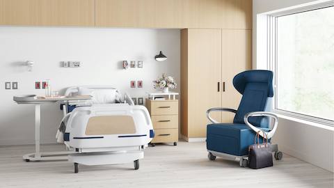 A patient room containing a hospital bed with a Nemschoff Bedside Cabinet next to it, a Mirage Overbed Table on the other side, and a Nemschoff Ava Recliner in blue upholstery nearby.