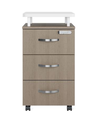 A Nemschoff Bedside Cabinet in a bleached beige finish with three drawers, champagne metallic arc pulls, a keyless lock, a raised top in white laminate, and casters.