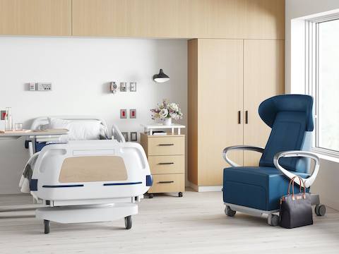 A patient room containing a hospital bed with a Nemschoff Bedside Cabinet next to it, a Mirage Overbed Table on the other side, and a Nemschoff Ava Recliner in blue upholstery nearby.