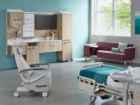 A patient room environment with a Compass footwall in an elm finish, a Nemschoff Ava patient recliner in a light gray upholstery, a Nemschoff Palisade Flop Sofa in a purple upholstery, and a patient bed with an overbed table.