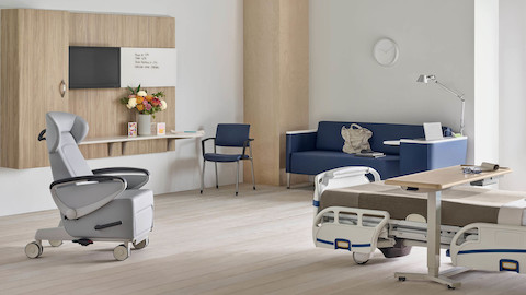 Compass System footwall in a patient room with a gray Nemschoff Ava patient recliner, a blue Nemschoff Palisade Flop Sofa, a fully-upholstered blue Verus side chair, and a Mirage Overbed Table over a hospital bed.
