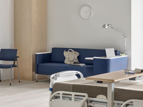 A patient room with a blue Nemschoff Palisade Flop Sofa, a fully-upholstered blue Verus side chair, and a Mirage Overbed Table over a hospital bed.