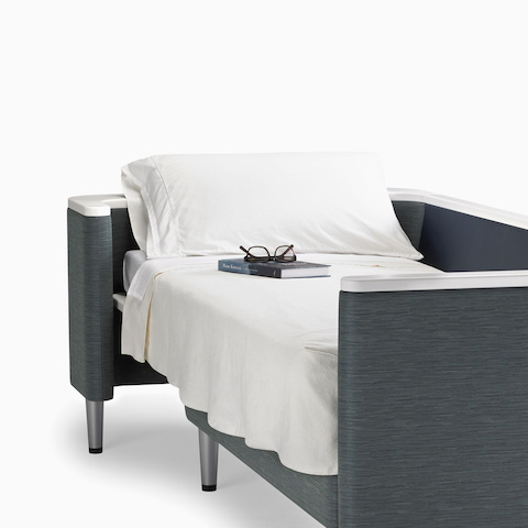 A Nemschoff Palisade Flop sofa converted to sleep surface in dark gray textile with white solid surface arms caps and white pillow and sheet.