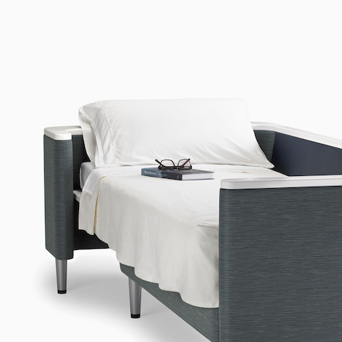 A Nemschoff Palisade Flop sofa converted to sleep surface in dark gray textile with white solid surface arms caps and white pillow and sheet. A Nemschoff Palisade Flop sofa converted to sleep surface in dark gray textile with white solid surface arms caps and white pillow and sheet.