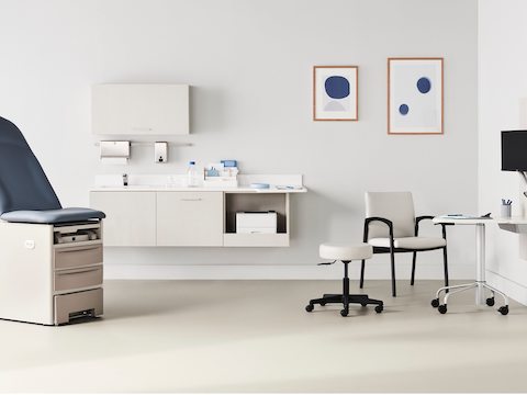Exam room with a Brewer Company white and blue exam table, light wood Mora casework, a light wood Intent Solution wall featuring a mobile, height-adjustable table, surrounded by a Valor Side Chair in a light gray upholstery and black base and a physician stool.