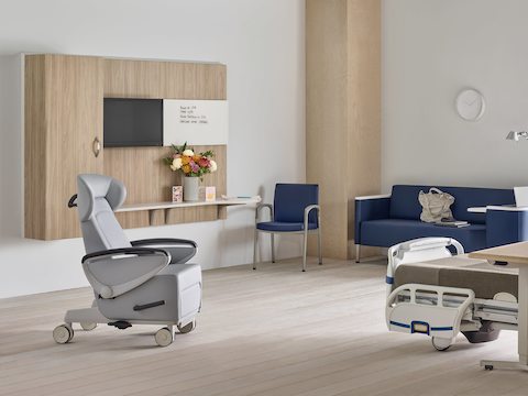 Compass System footwall in a patient room with a gray Ava patient recliner, a blue Palisade Flop Sofa, a blue Valor Side Chair, and a Mirage Overbed Table over a hospital bed.