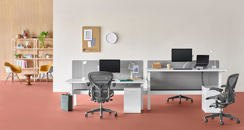 An office setting with a Nevi Link standing desk system in the foreground with white rectangular work surfaces and black Aeron office chairs, and a lounge area with Eames shell chairs and bookcase in the background. One of the four desks is raised to standing height.