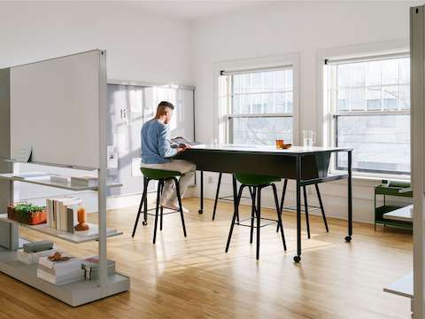 A man sitting at a black double sided OE1 Communal Table with casters, with OE1 Agile Walls in the foreground and OE1 Wall Rail and Board and OE1 Storage Trolley in the background.