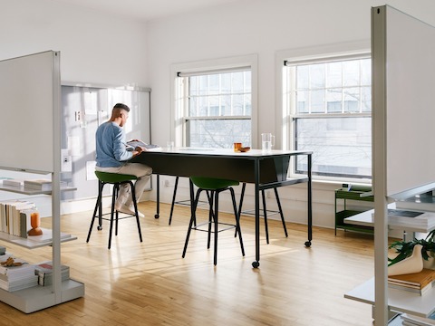 A man sitting at a black double sided OE1 Communal Table with casters, with OE1 Agile Walls in the foreground and OE1 Wall Rail and Board and OE1 Storage Trolley in the background.
