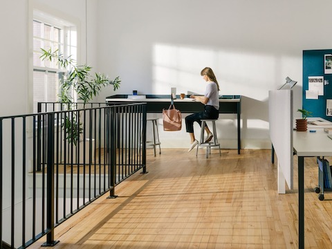 Mesas rectangulares OE1 en blanco en configuración de estaciones de trabajo, con una mujer sentada frente a una Mesa comunitaria OE1 en azul, un Atril móvil OE1 en azul y un tablero de proyectos al fondo.