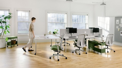 A person pushes an OE1 Project Table to four grey OE1 Rectangular Tables to form a bench, with green OE1 Storage Trolleys and green Lino Chairs, and an OE1 Wall Rail and Board in the background.