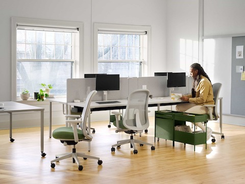 Una persona se sienta junto a una mesa rectangular OE1 en gris unida con otras mesas para formar una banca, con una Mesa de proyectos OE1 en gris y sillas Lino en verde en el frente y un Riel y panel para pared OE1 al fondo.