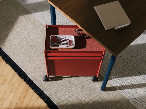 A red OE1 Storage Trolley with casters, nesting under an OE1 Rectangular Table with dark brown surface and blue legs.