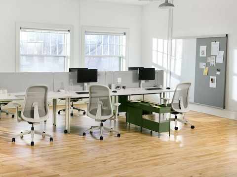 Cuatro mesas rectangulares OE1 en gris y una Mesa de proyectos OE1 en gris juntas para formar una banca, con Carritos de almacenamiento OE1 en verde, sillas Lino en verde y un Riel y panel para pared OE1 al fondo.