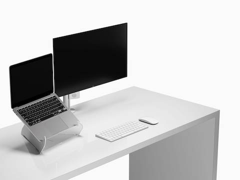 Vista em ângulo de um laptop aberto sobre um suporte para laptop Oripura, ao lado de um braço para monitor e uma tela em cima de uma superfície de trabalho com ferramentas de trabalho