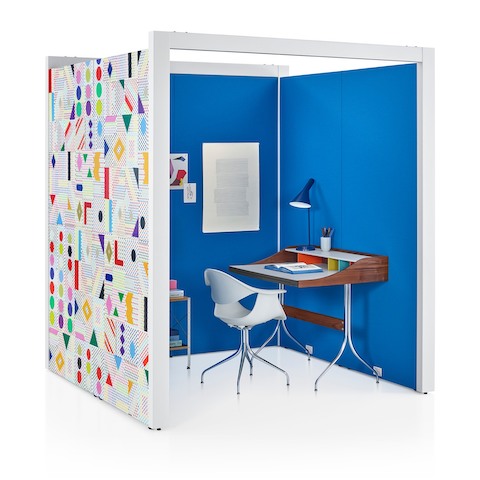 Un espacio Overlay de tres lados con interiores de tejido azul apto para pinchar notas y patrón geométrico colorido en la pared exterior con una silla y escritorio en su interior.