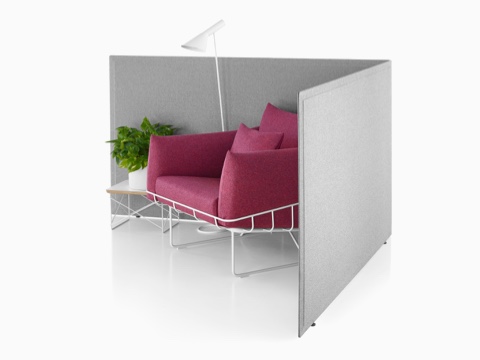 Viewed at an angle, two gray fabric Pari freestanding privacy screens connected in a L-shape configuration with a settee, end table, and lamp in it.