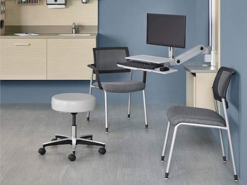A patient room featuring a Physician Stool in gray fabric, pulled up to a computer work surface. Mora System and Verus Side Chairs are featured nearby.