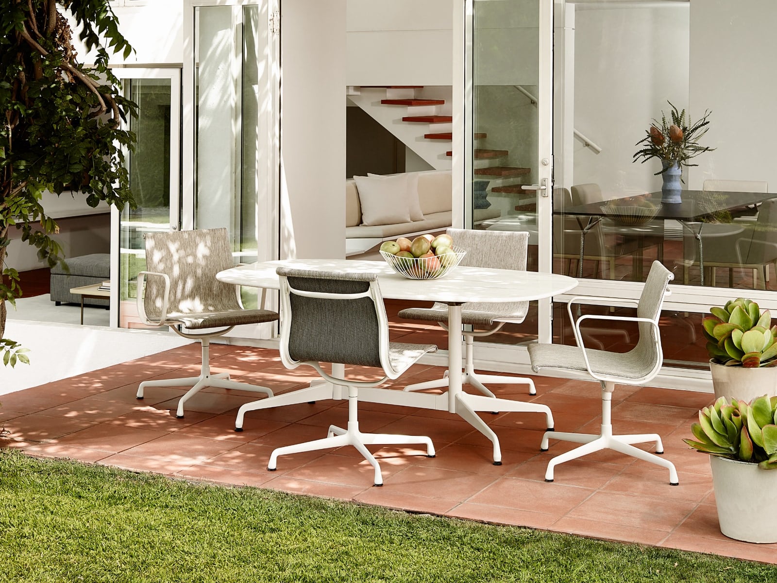 Quatro cadeiras para visitantes externas Eames Aluminum Group cercam uma mesa externa Eames em um pátio.