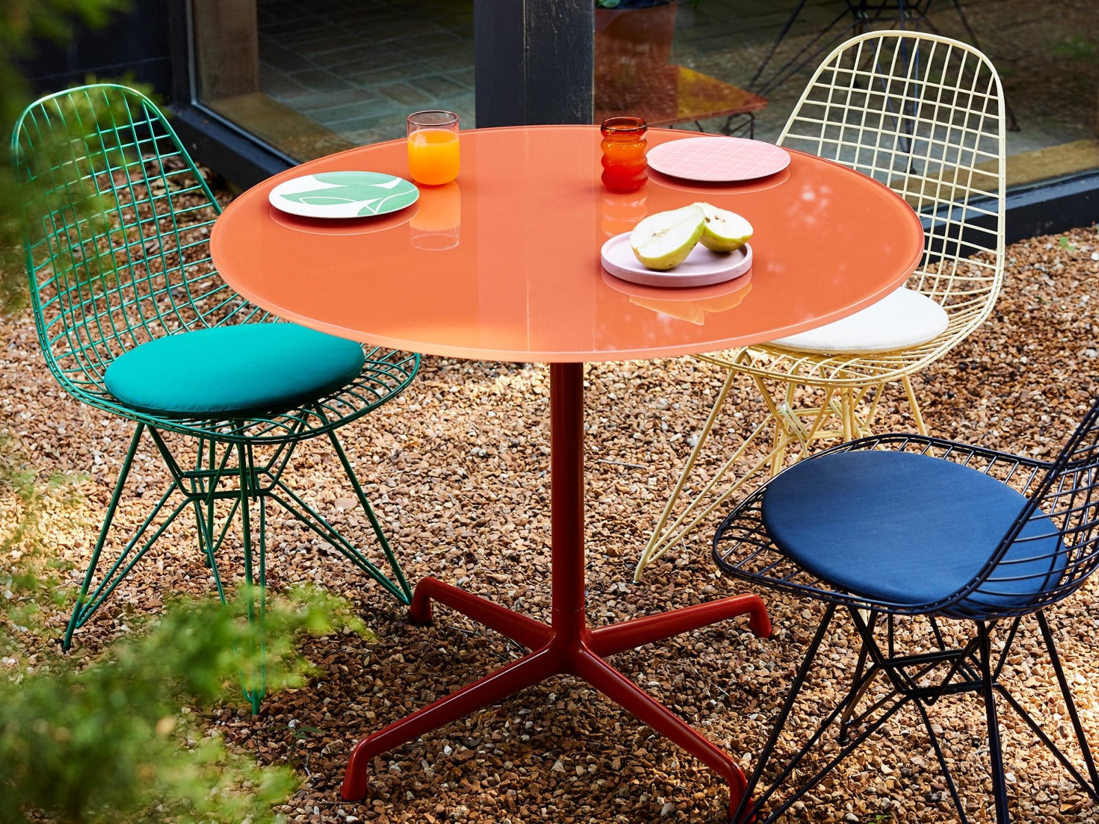 Un ambiente exterior de Herman Miller x HAY; sillas Eames Wire verdes, azul oscuro y amarillas para exterior y una mesa Eames roja de hierro para exterior.