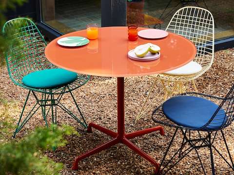 An outdoor setting of Herman Miller x HAY; green, black blue, and yellow Eames Wire Chairs-Outdoor; and an iron red Eames Table-Outdoor.