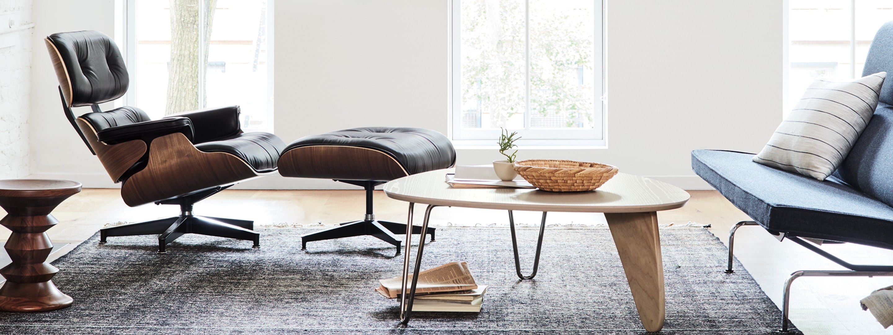 客厅摆放着 Eames 休闲椅和脚凳、Noguchi Rudder 桌子、Eames 紧凑型沙发和 Eames 胡桃木凳子，背景为大窗户。