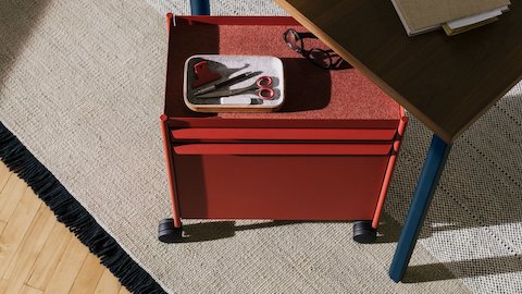 Un Carrito de almacenamiento OE1 en rojo con ruedas giratorias, encajado debajo de una mesa rectangular OE1 con superficie en marrón oscuro y patas en azul.