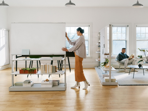 A woman moving a grey OE1 Agile Wall with a man sitting on a checkerboard Nelson Coconut Lounge Chair in the background. A woman moving a grey OE1 Agile Wall with a man sitting on a checkerboard Nelson Coconut Lounge Chair in the background.