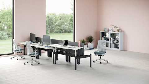 A bank of six Ratio desks with one positioned at a standing height. Three green and white Verus Chairs are set around the desks with a grey CK2 storage unit in the background.