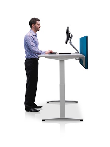 Mesa com ajuste de altura Ratio posicionada a uma altura em pé com com uma tela de privacidade azul.