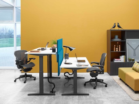 Mesas com ajuste de altura Ratio uma de frente para a outra, posicionadas nas alturas para trabalhar sentado e em pé, combinadas com cadeiras de escritório Aeron pretas.