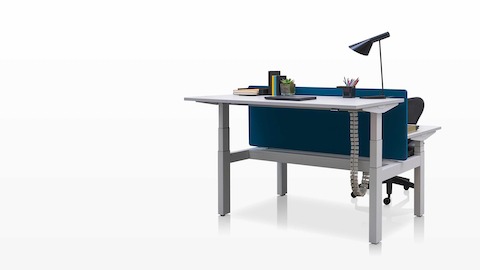 Mesas com ajuste de altura Ratio, posicionadas uma de frente para a outra em alturas para trabalhar sentado e em pé e separadas por uma tela de privacidade azul.