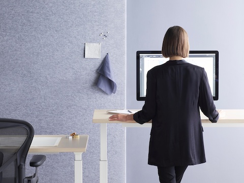 A workspace with a woman using a rectangular Renew Sit-to-Stand Table at a standing height with another Renew Table and an Aeron Chair beside her.