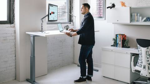 A man works on a computer while standing at a rectangular Renew Sit-to-Stand Table.