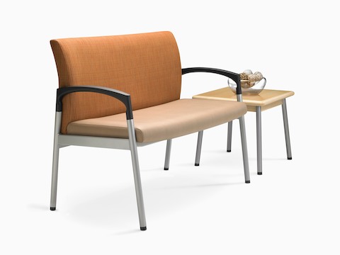 带角度的视图：配有橙色靠背和米黄色椅座的加宽款Valor Plus座椅，摆放在方形茶几旁边。
