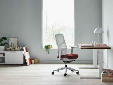 A Verus Chair with a dark red upholstered seat and white Triflex back next to a Renew Sit-to-Stand Table in a home office with gray walls and light hardwood floors. A Verus Chair with a dark red upholstered seat and white Triflex back next to a Renew Sit-to-Stand Table in a home office with gray walls and light hardwood floors.