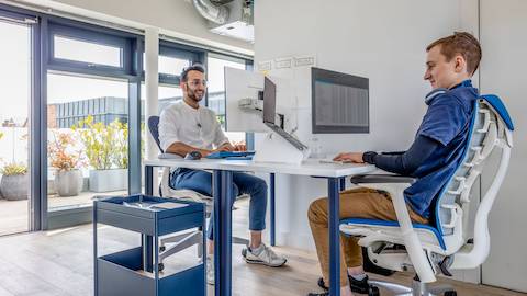Twee mannen werken aan OE1 tafels met Oripura laptophouders. Twee mannen werken aan OE1 tafels met Oripura laptophouders.