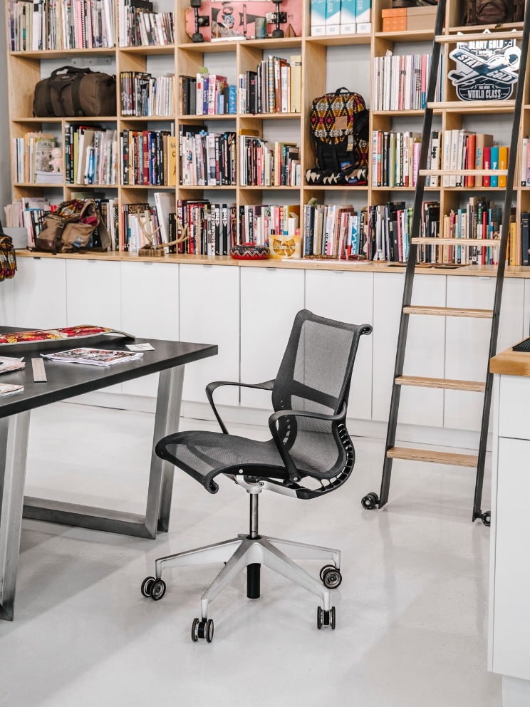 A Setu chair sits empty by a desk, with a large bookcase and ladder in the background. A Setu chair sits empty by a desk, with a large bookcase and ladder in the background.
