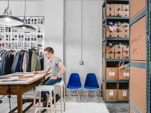 A man works at a wooden desk, seated on a Magis stool with two blue Eames chairs nearby.