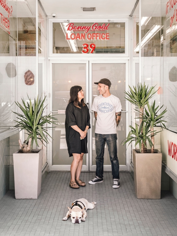 Two people converse in front of office doors as their dog lies on the ground. 