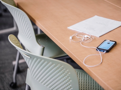 A smartphone with earpieces sits on a desk net to a stack of papers. 