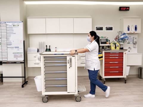 戴着口罩、穿着白色和蓝色手术服的护士推着一辆Compass系统供应推车沿着走廊行走。
