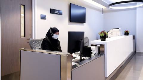 Una mujer con máscara facial sentada en una silla Mirra 2 en gris claro en la estación de enfermería en gris y blanco. Hay puertas de madera y una gran pantalla de TV detrás de la enfermera.