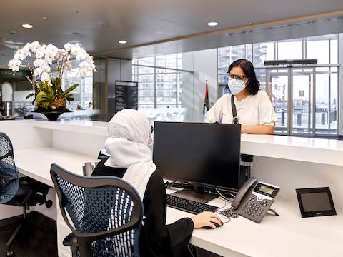 Recepcionista vista de atrás sentada en una silla Mirra 2 en gris y azul, frente a su computadora, mientras conversa con otra mujer en la recepción del hospital.