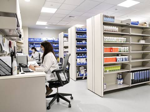 Two ladies in an apothecary space sitting on a grey Mirra 2 chairs and working at their desks, on the right shelving units with medication and files in various colors.