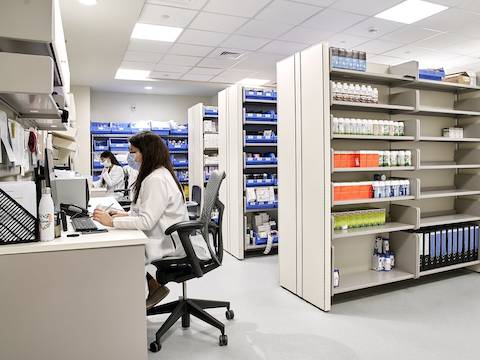 Dos mujeres en un espacio de boticario sentadas en sillas Mirra 2 en gris, mientras trabajan en sus escritorios. Sobre la derecha, hay estanterías con medicamentos y archivos en distintos colores.