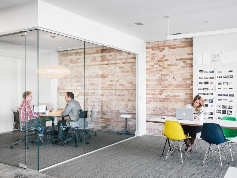 Two people talk in a glass-walled conference area, while a woman works on a laptop. 