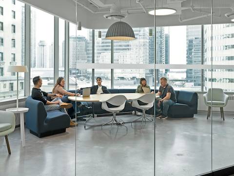 Five people sit on blue Chadwick Modular Seating in a glass-walled conference room. Two white Nelson Swag Leg chairs sit across the table from them, and a gray Saiba Chair sits off to the side. 